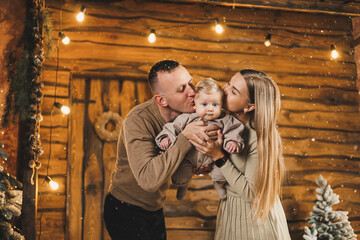 A caring couple is sitting near a beautiful Christmas tree and enjoying their son in stylish knitted clothes. Happy family moments during winter holidays.