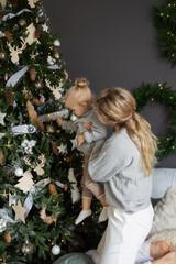 mother holds her young daughter decorate Christmas tree together. tree ornaments, lights, and ribbons. while woman gray sweater and white pants. concept: family christmas home family