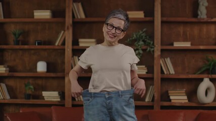 Happy senior woman holding large jeans after weight loss, standing in a cozy room.