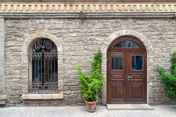 A stone building features a wooden door beside a decorative wrought iron window, with potted greenery enhancing the entrance.