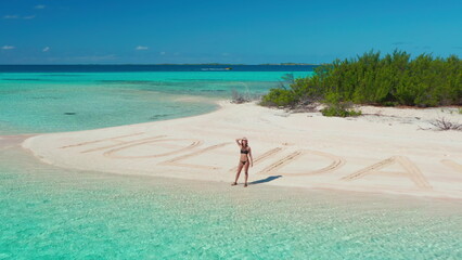 Obraz premium Young woman standing alone on a tropical beach with holidays written in the sand, surrounded by turquoise waters, enjoying the warm sun and feeling free and adventurous
