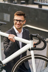 Obraz premium A smiling businessman in a suit, carrying a white bicycle confidently in an urban setting.