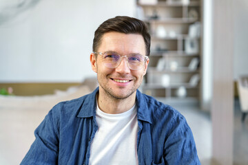 Obraz premium A smiling man in glasses and a blue shirt sits indoors, creating a warm and inviting atmosphere.