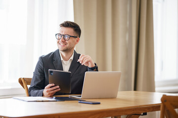 cheerful businessman in a suit works with a tablet and laptop in a bright, modern office.