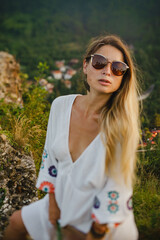 A girl with long blonde hair in a hat, boots and a white long dress on the background of a panorama of green mountains, Alps in Europe at sunset. A girl with a bouquet of lavender flowers, wearing sun