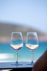 Two partially filled wine glasses stand on a table with an ocean and a blurred mountain backdrop, capturing the essence of relaxation and scenic beauty on a summer day.