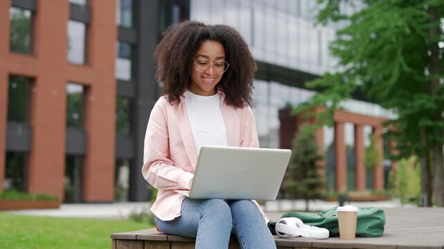 African american female student works typing on laptop sitting on a bench in campus space near university building. Young black woman in glasses studies remotely, prepares homework, completes tasks