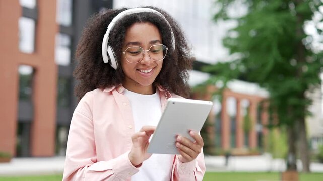African american female student using digital tablet for learning sitting on bench on street near university building. Young black woman in wireless headphones chats online or browses web. Close up