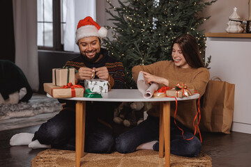 positive young millennials floor Christmas tree wrapping presents together. man wearing Santa hat....