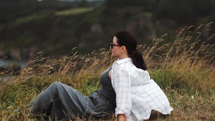 Romantic, lyrical mood. A girl sitting in the grass and flowers on a cliff near the ocean coast, with a beautiful view of rocks and cliffs, strong wind and comfortable clothes, meditation and peace