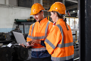 Two engineers are wearing orange safety vests and hard hats are looking at a laptop at locomotive...