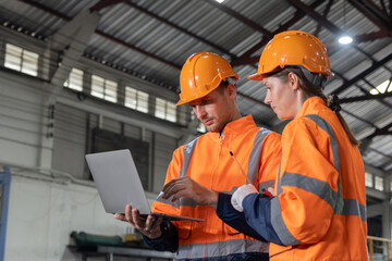 Two engineers are wearing orange safety vests and hard hats are looking at a laptop at locomotive service station.