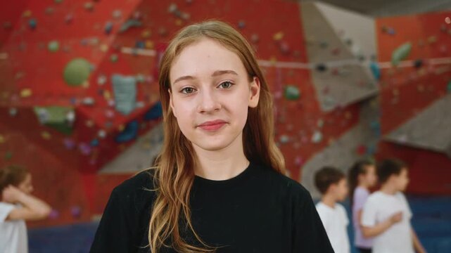 Portrait, a pretty and sweet blonde girl of 14 years old stands in front of the camera in the gym inside the climbing wall. Friends are playing or messing around in the background, running on gym mats