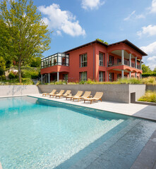 Red-walled villa in a big green garden with trees. There is a big swimming pool and sun loungers. Sunny summer day.