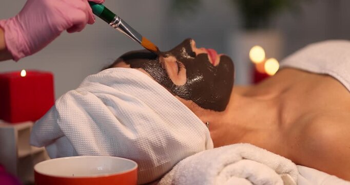 Young woman applies detoxifying clay mask to face in beauty salon. Facial skin care and rejuvenation concept