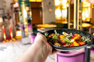 A close-up of a hand holding a plate of fresh salad with various vegetables, set against a blurred restaurant background. Ideal for concepts of healthy eating, dining out, and lifestyle.