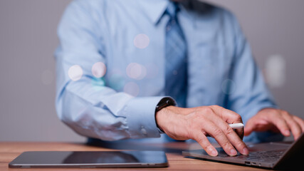 A man in a blue shirt and tie is typing on a laptop. He is using a pen to write on a tablet. Concept of professionalism and focus, as the man is working on a task that requires concentration