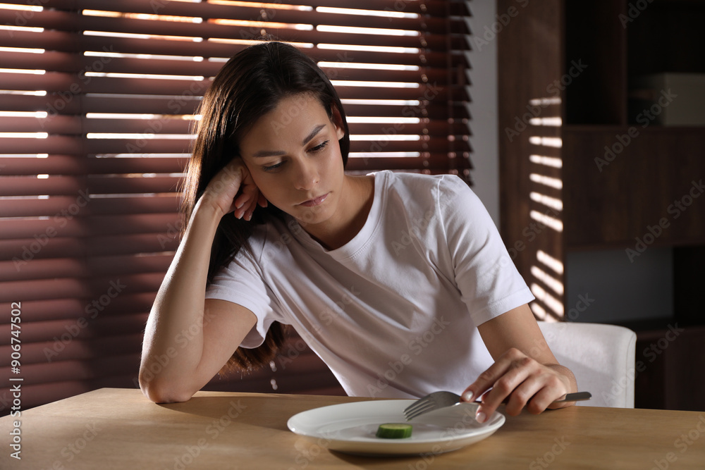 Wall mural Eating disorder. Sad woman with fork and slice of cucumber at wooden table