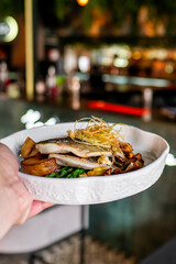 A close-up of a gourmet dish featuring seared fish on a bed of green vegetables and mushrooms, garnished with herbs. The dish is held in hand, showcasing its vibrant colors and fresh ingredients.