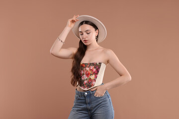 Beautiful woman in stylish corset and hat posing on beige background