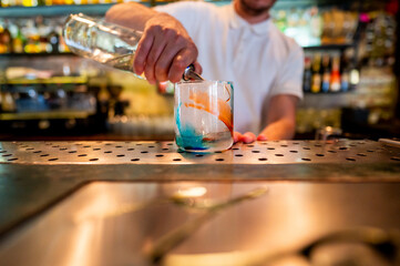 bartender’s hand pours a clear liquid into a cocktail glass with blue swirls, set on a bar counter with various tools nearby. The scene captures the art of mixology in stylish and modern bar setting.