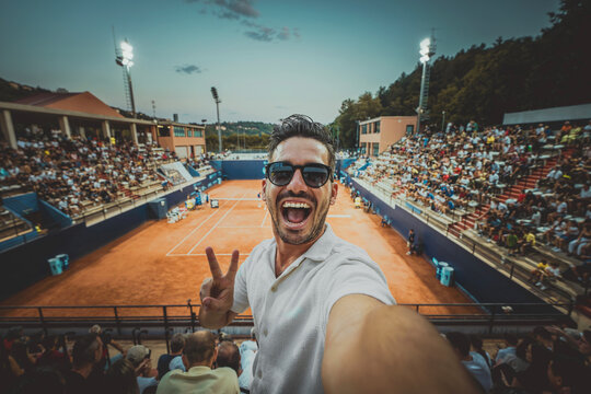 Handsome young tennis spectator taking selfie picture watching grand slam tournament match at the stadium - Sport and technology life style concept - Powered by Adobe