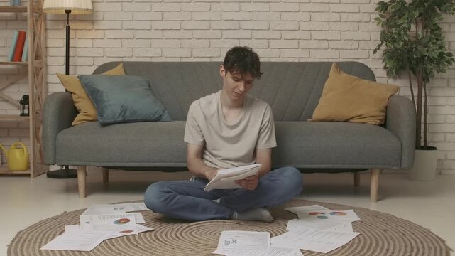 Focused adult man college student sitting on the floor in apartment room at daylight and doing homework writing in notebook. Education concept.