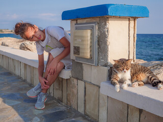 Smiling girl looking and peeking at a non-pedigree tabby striped cat with closed eyes having rest and relaxing at the sea promenade in Ayia Napa.