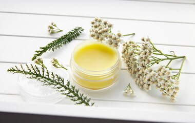 Balm made with Achillea millefolium, yarrow or common yarrow. Jar with medicinal balm on white wood background. Fresh flower for decoration. Still life. © FotoHelin