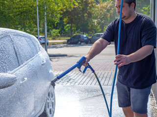Man with the high powered hose in his hand covers with foam and washes his car at a self-serve car wash. Avoid personal contact and wash alone.