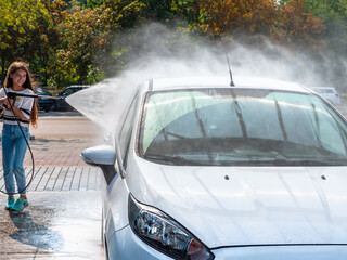 Girl teenager with the high powered hose in her hands washes the car at a self-serve car wash. Small water drops around. Avoid personal contact and wash your car alone.