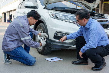 Asian male insurance officer in uniform crouching by vehicle and using smartphone to document bumper damage while client points at specific area. Emphasizes detailed inspection, customer communication