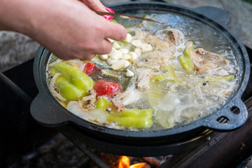 The woman's hands add vegetables to the cauldron, cooking over an open fire.