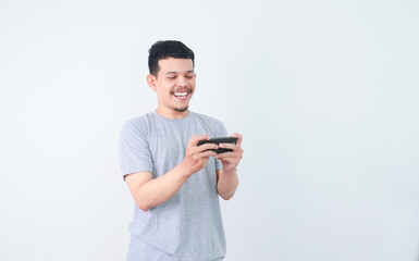 Portrait of an Indonesian Asian man, wearing a grey T-shirt, expressively playing a game on his smartphone, isolated against a white background.