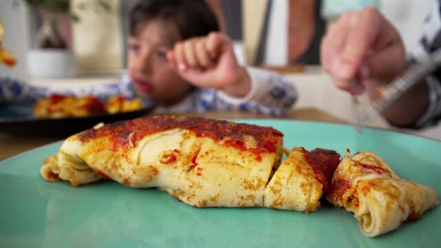 Parent cutting a serving of homemade enchiladas on a plate while a child sits in the background during a family mealtime