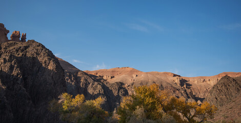 autumn leaves contrast with the stark, imposing cliffs, as the soft glow of the setting sun bathes the serene desert landscape in a warm, golden light.