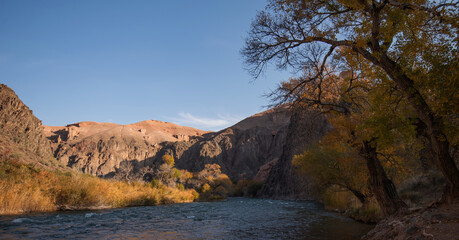 Amber foliage frames a mountains river, meandering at the foot of towering, rugged cliffs under the gentle embrace of a clear azure sky.