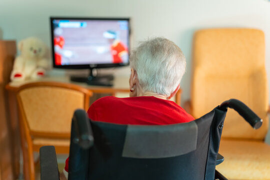 Senior woman with her back turned sitting in a wheelchair while enjoying watching TV in solitude at home
