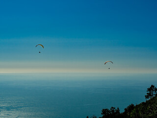 two paraglider sails soar through the air against a very blue sky and blue sea to create a scene of...