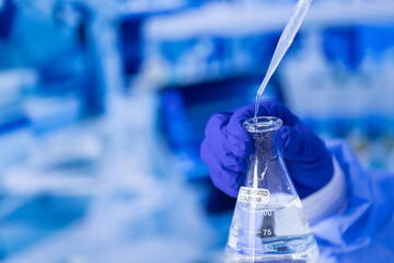 A person is holding a beaker with a liquid in it. The liquid is clear and the beaker is labeled with the word 