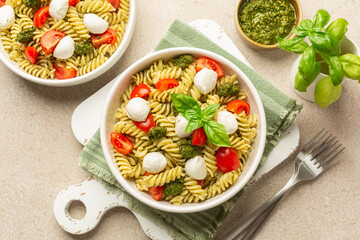 Cold pasta salad with homemade pesto sauce, tomato and mozzarella cheese, decorated with basil leaves on a beige table surface. Italian lunch.