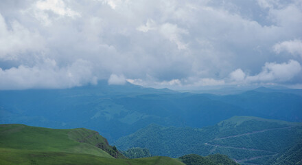 a tranquil panorama, rolling hills and gentle slopes are clad in vibrant green, with distant mountains partially veiled by a soft mist. Above, a dramatic sky, where billowing clouds