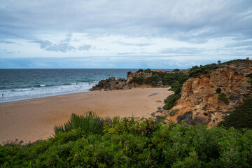 A secluded sandy beach bordered by rugged cliffs and Mediterranean shrubbery, with a solitary figure walking by the water under a partly cloudy sky.