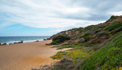 A secluded sandy beach bordered by rugged cliffs and Mediterranean shrubbery, with a solitary figure walking by the water under a partly cloudy sky.