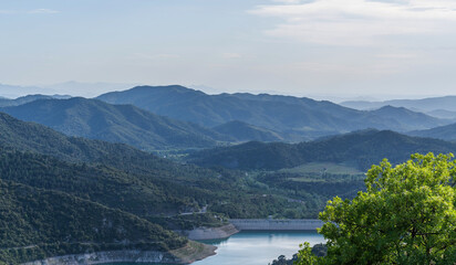 The serene landscape showcases undulating mountains with a dam's gentle curve nestled in the valley, framed by a foreground of vibrant green foliage.