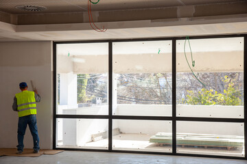 Construction worker smoothing drywall near a large window, showcasing expertise and attention to...