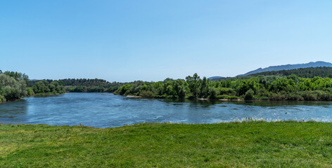 A tranquil river cuts through lush foliage under an expansive blue sky, with distant hills rolling into the horizon, embodying serene natural beauty.