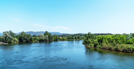 Overlooking a serene river, the foreground showcases a rustic terracotta-tiled roof, with lush greenery on the banks and distant mountains under a clear expansive sky.