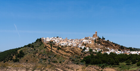 a Spanish hilltop village, its white buildings crowned with a church, perched atop a rugged hill, under the vast expanse of a clear blue sky.