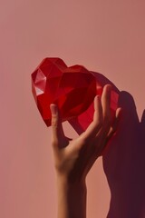 Red volumetric heart crafted from paper casts a hard shadow over a hand, against a pink background